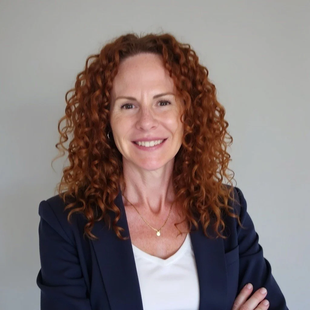 A woman with red curly hair smiling, wearing a navy blazer and a white top, standing against a light gray background.