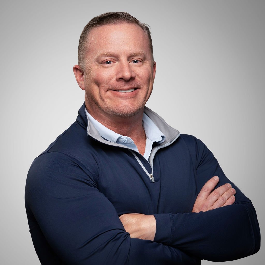 A smiling man with short, light-colored hair, wearing a dark blue quarter-zip jacket and a white shirt underneath, standing against a plain gray background with arms crossed.