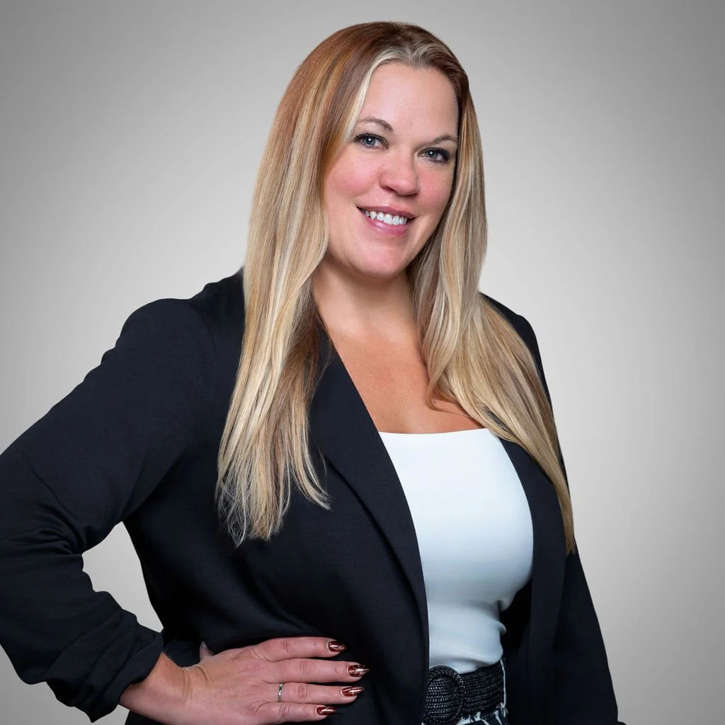 A blonde woman smiling, dressed in a black blazer, white top, and patterned pants, standing against a light gray background.