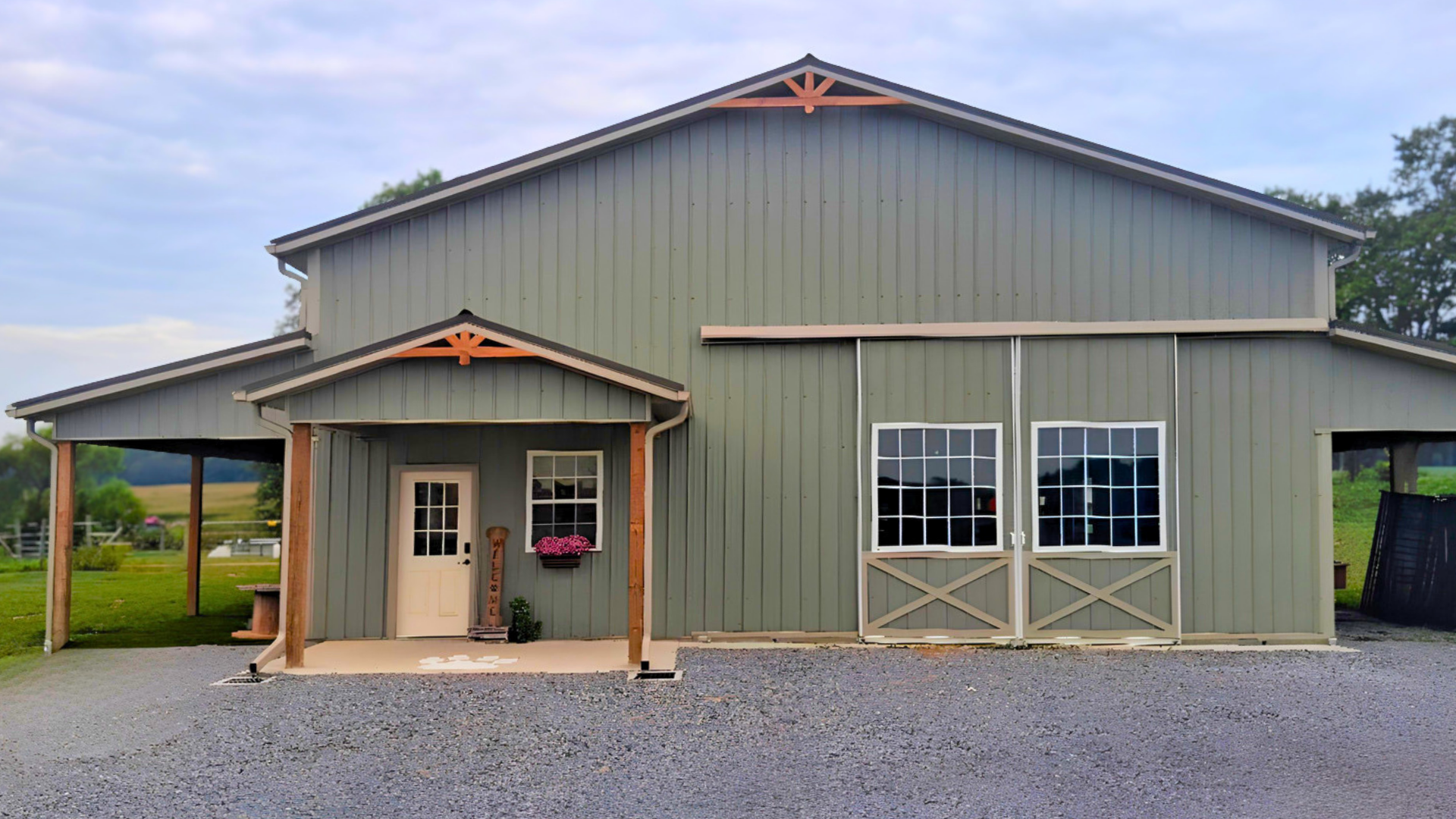 A large green farmhouse-style barn with two stories, a covered front porch, and a pair of large sliding doors. The barn has decorative wood trusses and is set on a gravel driveway with green fields and trees in the background.