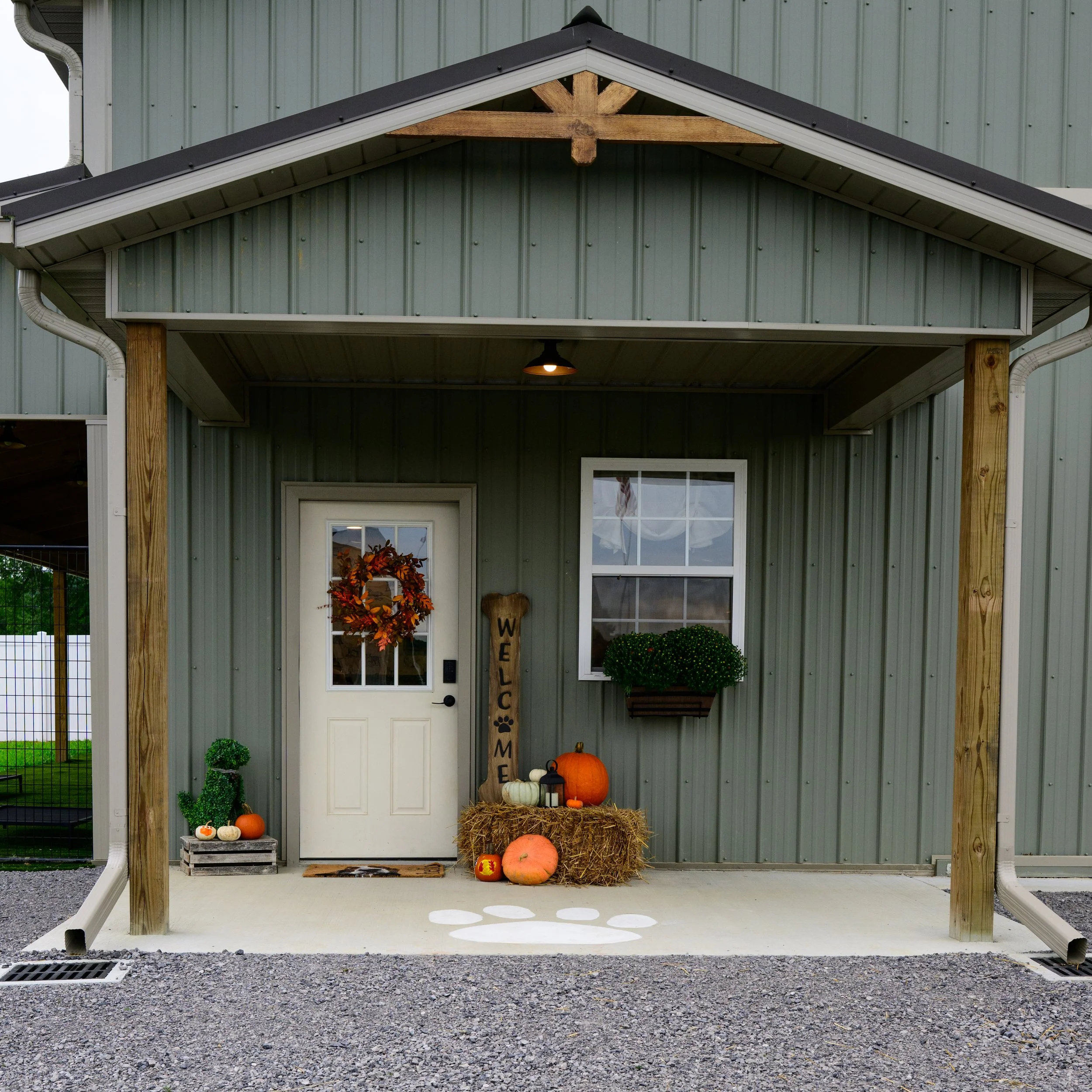 Front porch with fall themed decorations, including pumpkins, gourds, a harvest wreath, hay bales, a wooden welcome sign, and a potted plant, in front of a house with green siding and a white door.