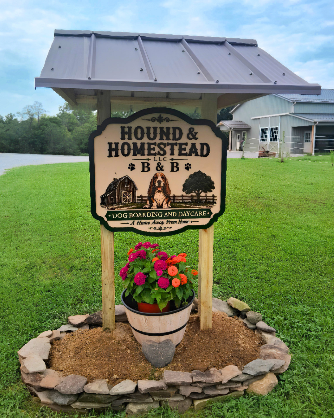 Sign for Hound & Homestead LLC B&B with a dog illustration, barn, tree, and a flower pot with colorful flowers in front, green lawn and house in background.