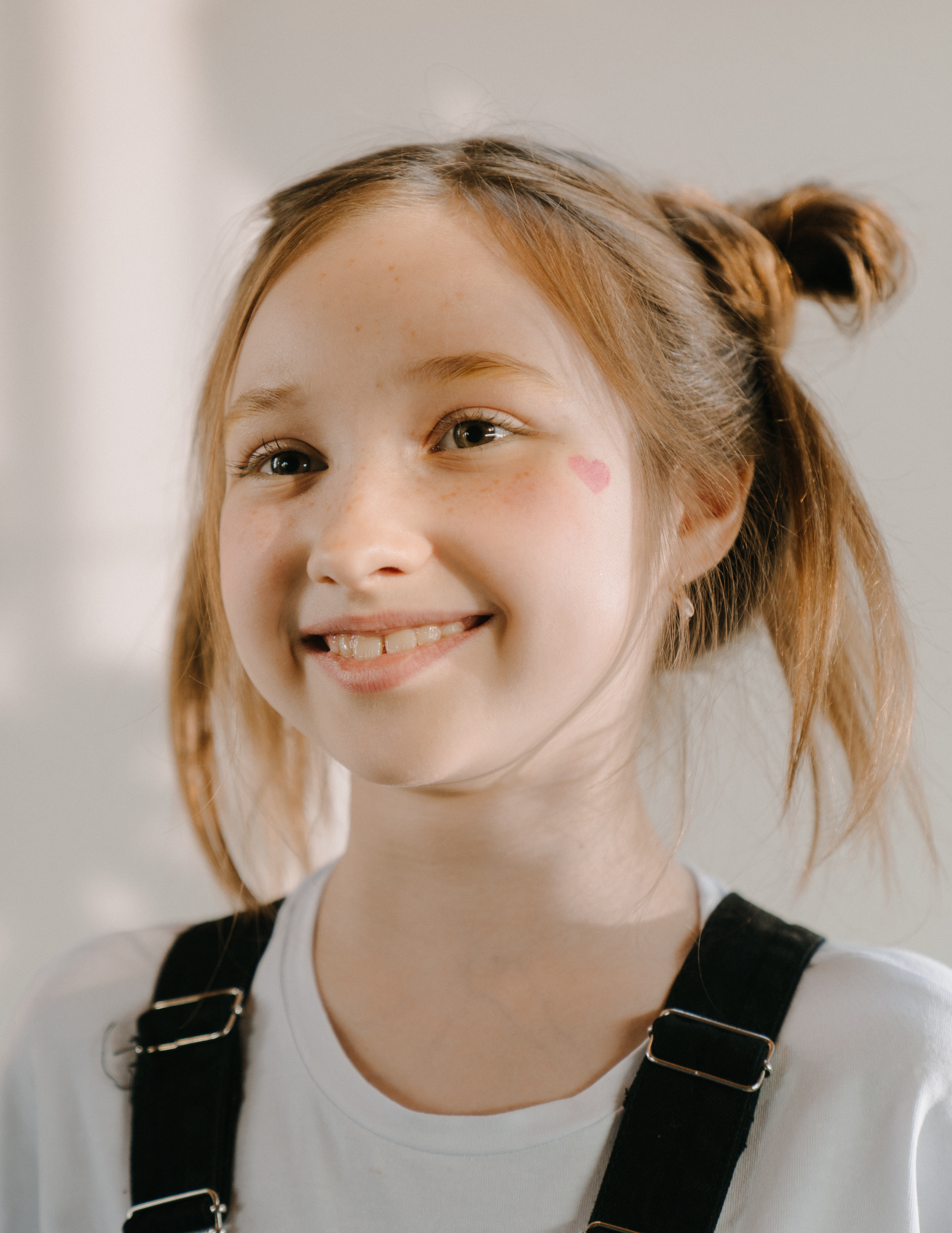 A young girl with red hair tied in a bun, smiling with a small pink heart sticker on her cheek, wearing a white shirt and black overalls.