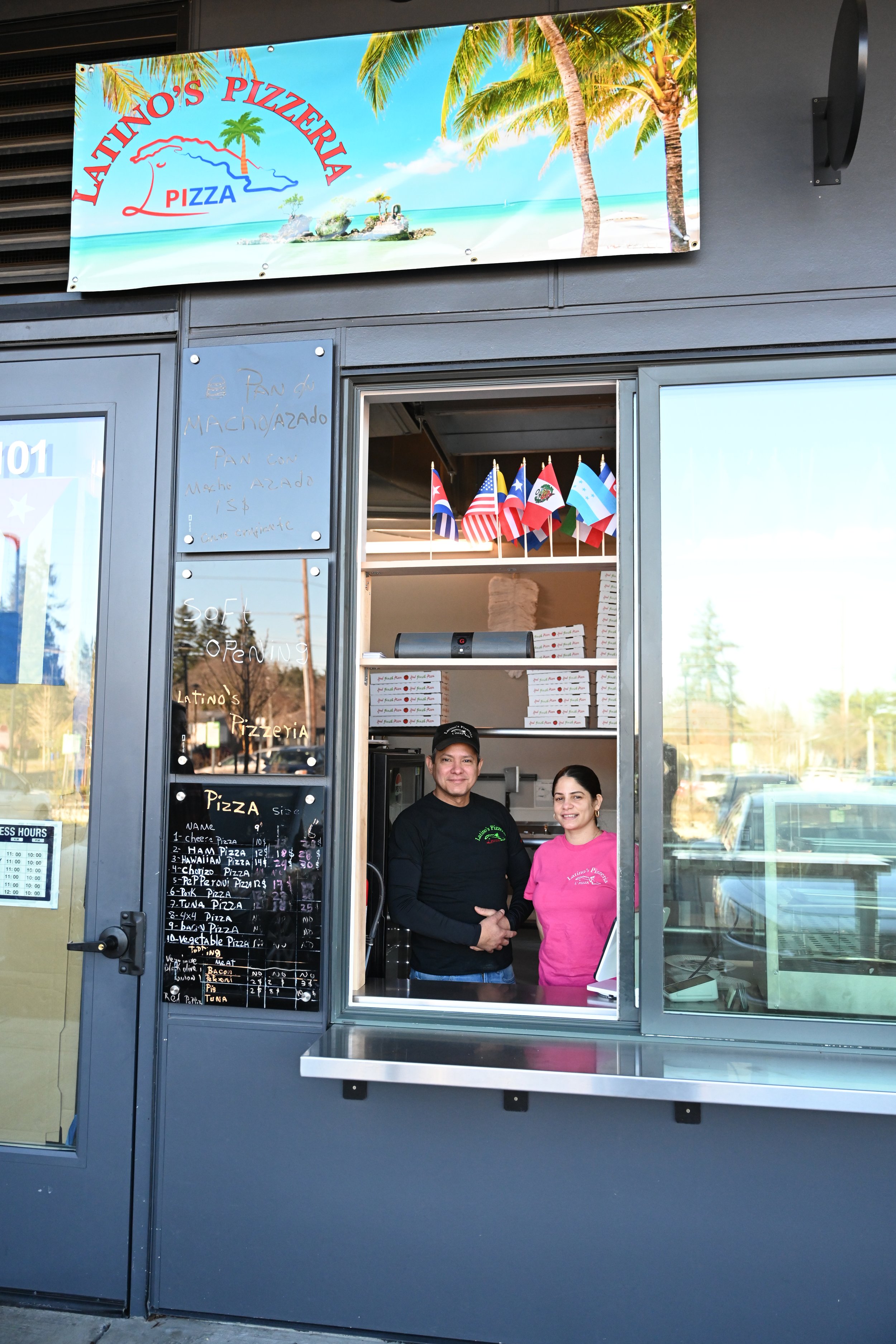 Owners of Latinos Pizzeria food stand with two employees smiling through a window, small flags hanging above, and a sign with menu items on the left.