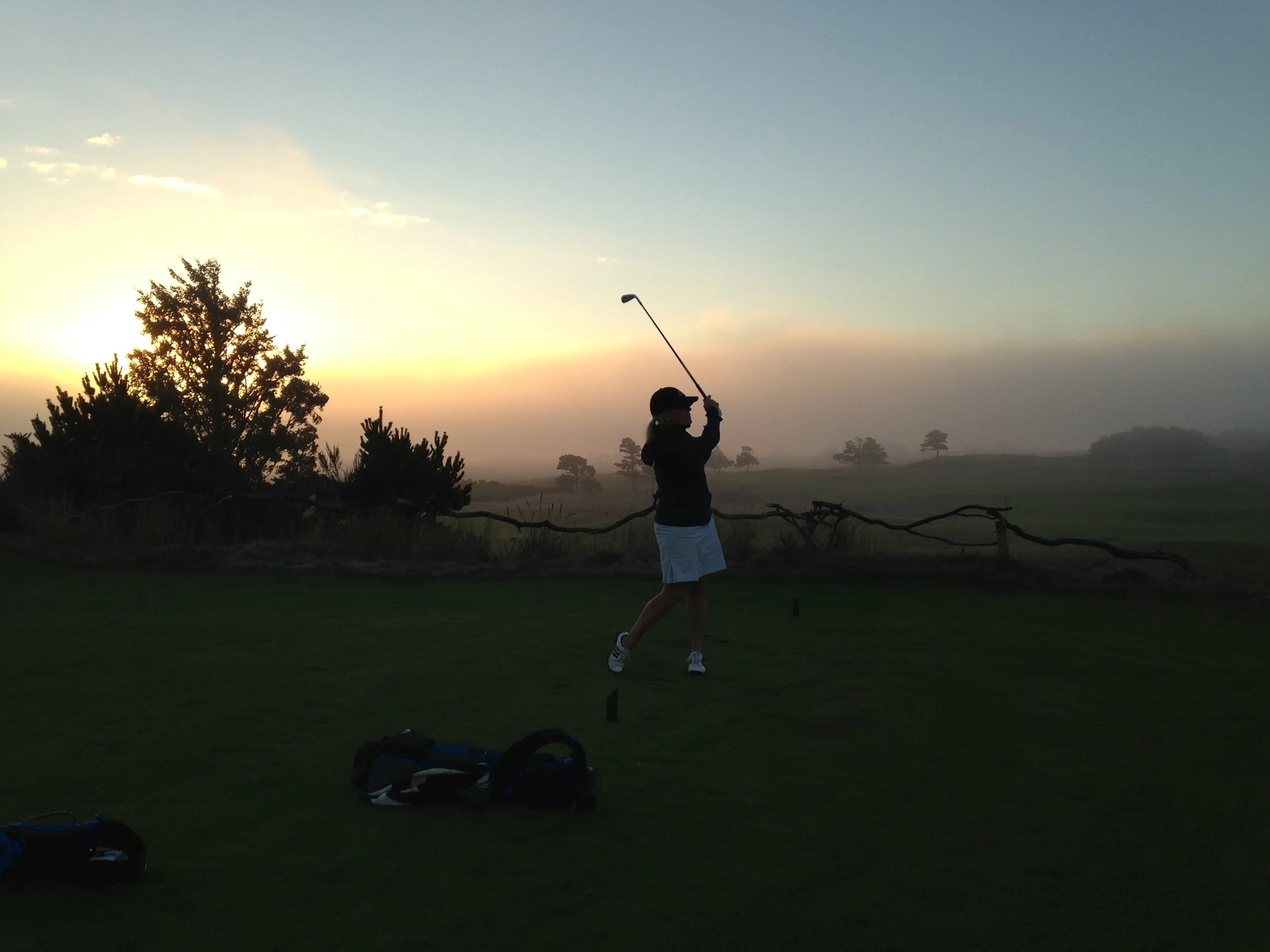 Person playing golf at sunrise on a golf course with trees in the background, a golf bag and equipment on the ground.