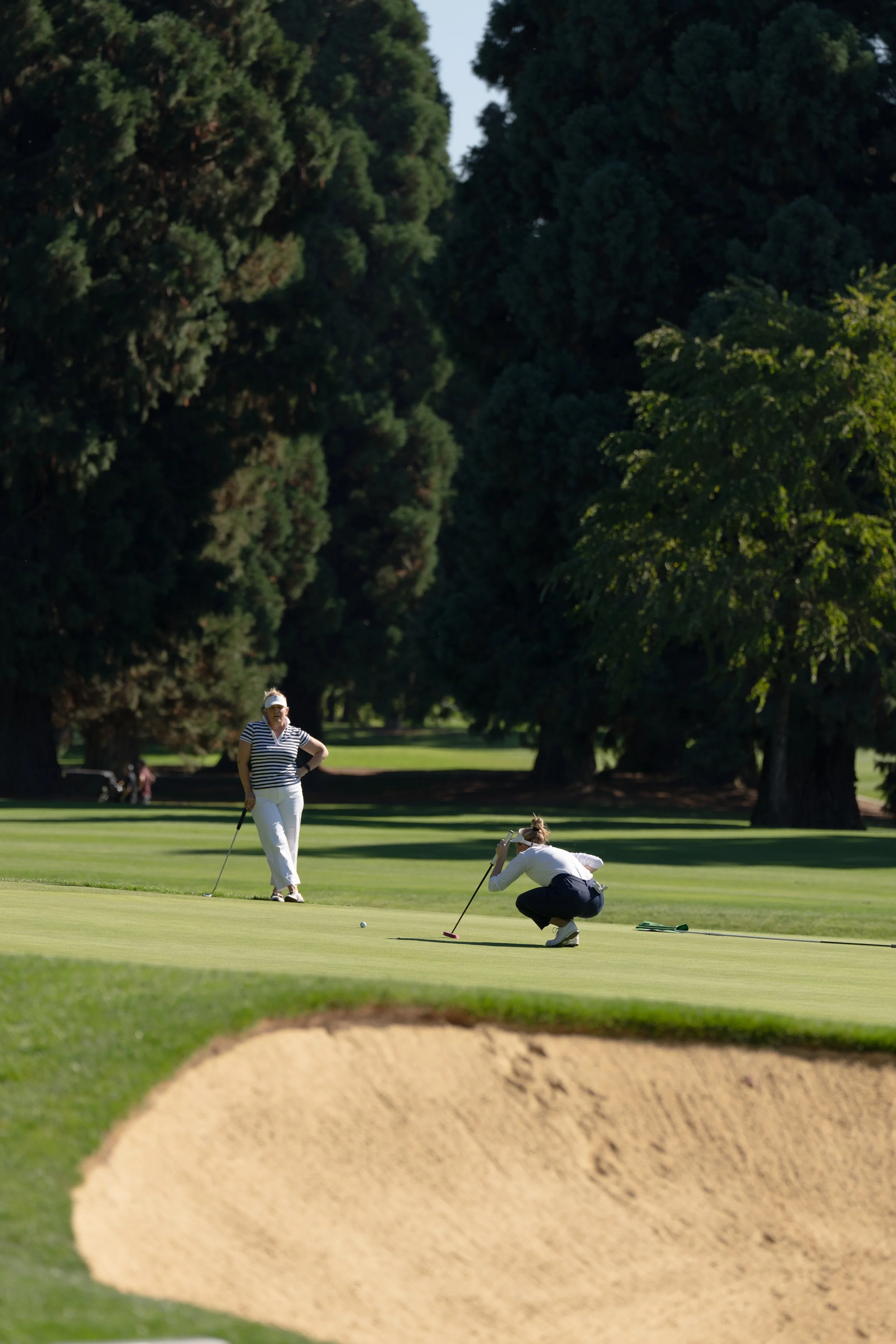 Two women playing golf on a golf course surrounded by large trees, with one woman putting and the other standing nearby.