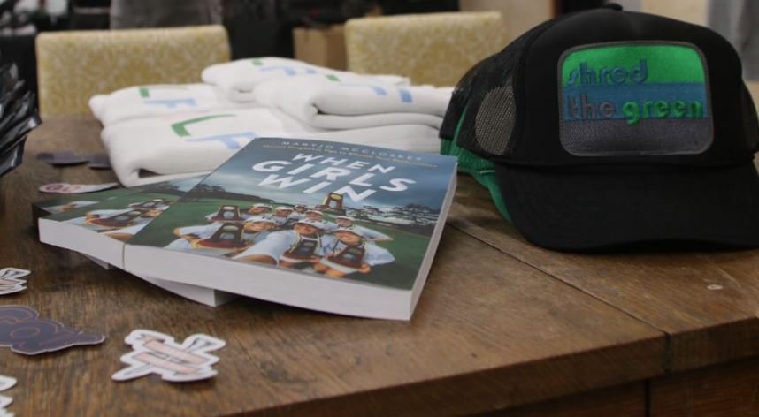 Books titled "When Girls Win" and merchandise including white shirts and a black baseball cap with text, on a wooden table.