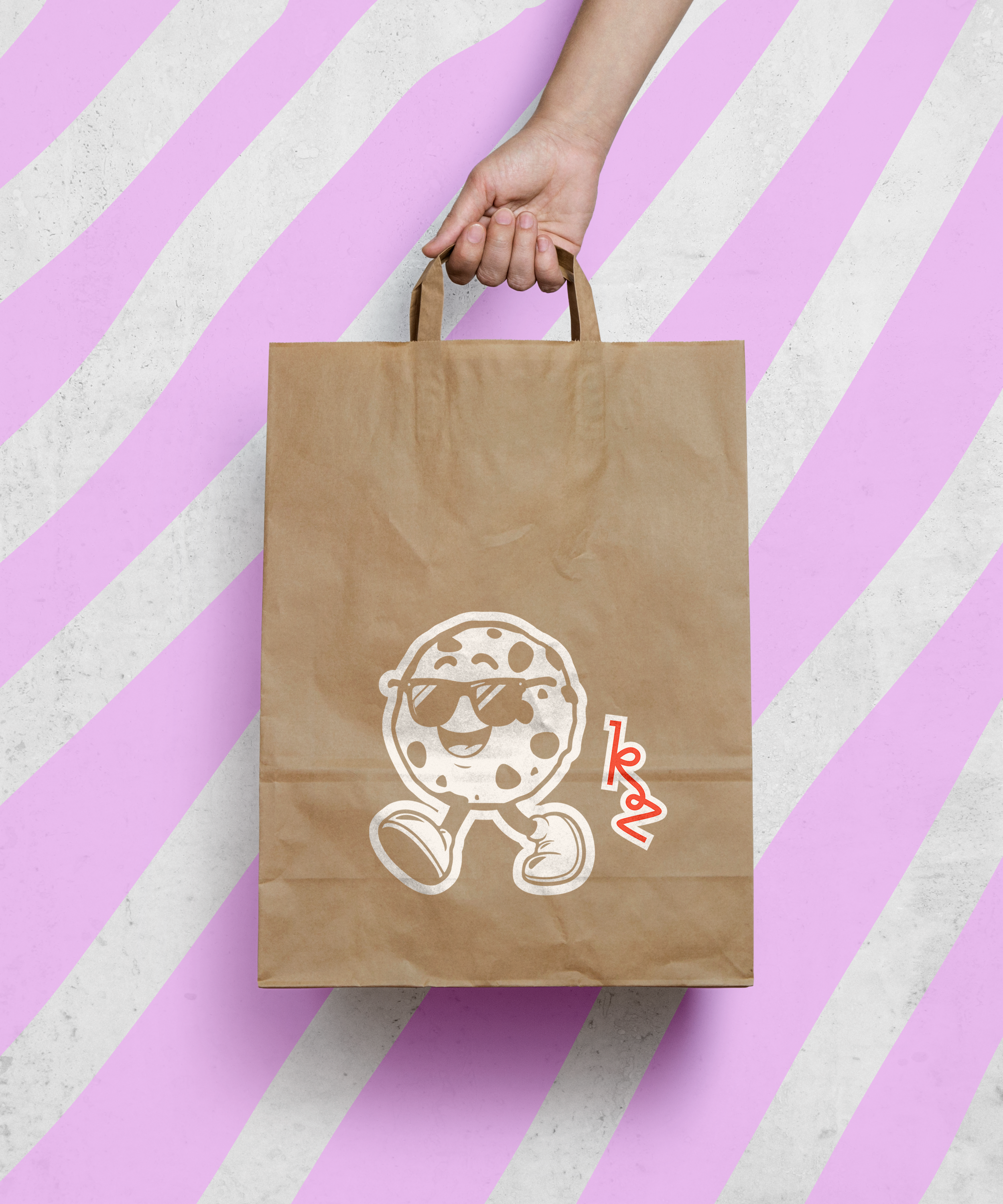 A craft paper bag with a smiling cookie mascot wearing sunglasses and sneakers printed on it, placed on a surface with purple and white diagonal stripes.