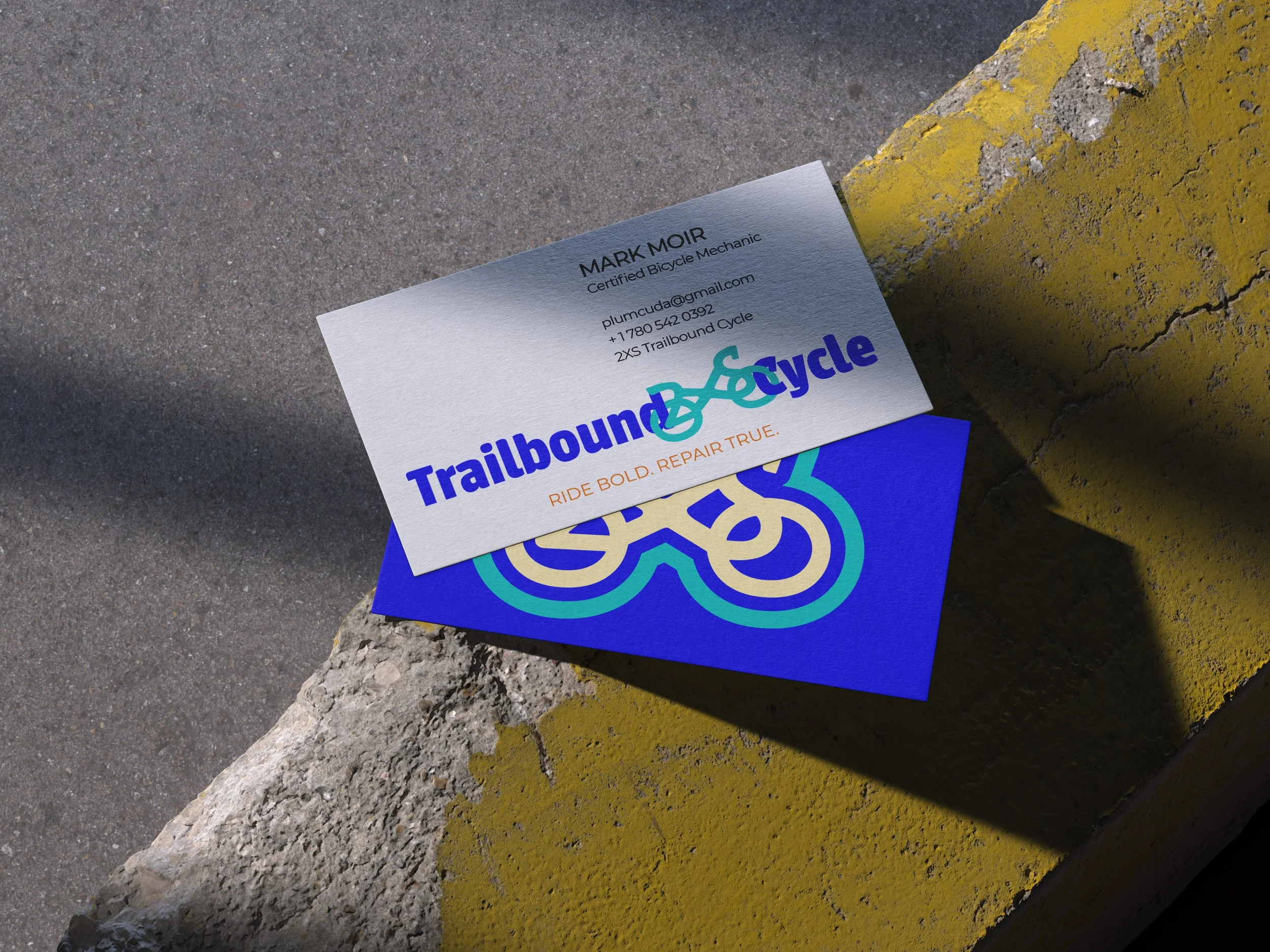 Business card for Mark Moir, a certified bicycle mechanic, placed on a concrete surface with yellow-painted curb and a large rock. The card features the Trailbound Cycle logo and contact information including email, phone number, and bicycle shop name.