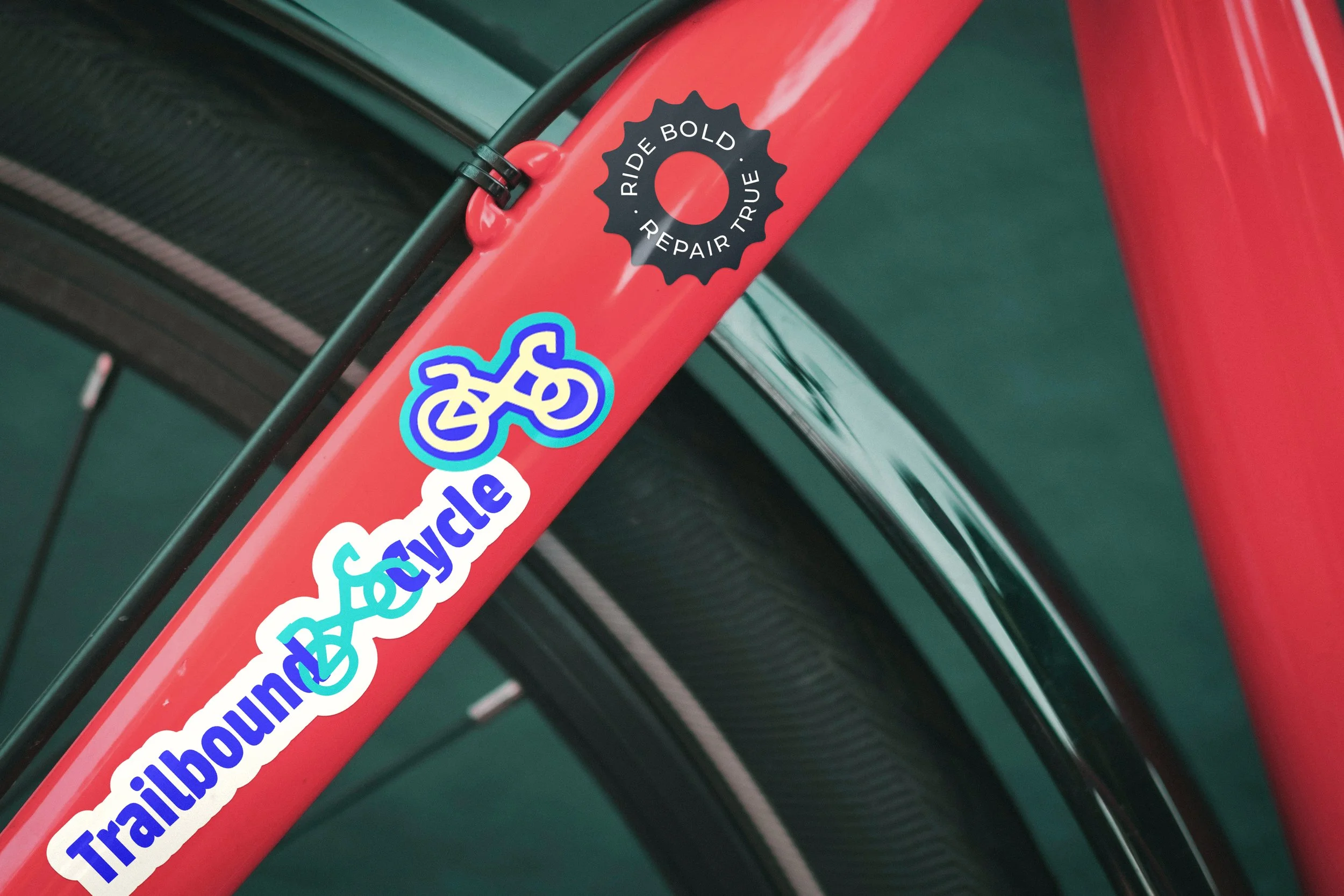 Close-up of a red bicycle frame with multiple colorful stickers, including a black circle sticker with the words 'RIDE BOLD. REPAIR TRUE' and a blue and white bicycle sticker.