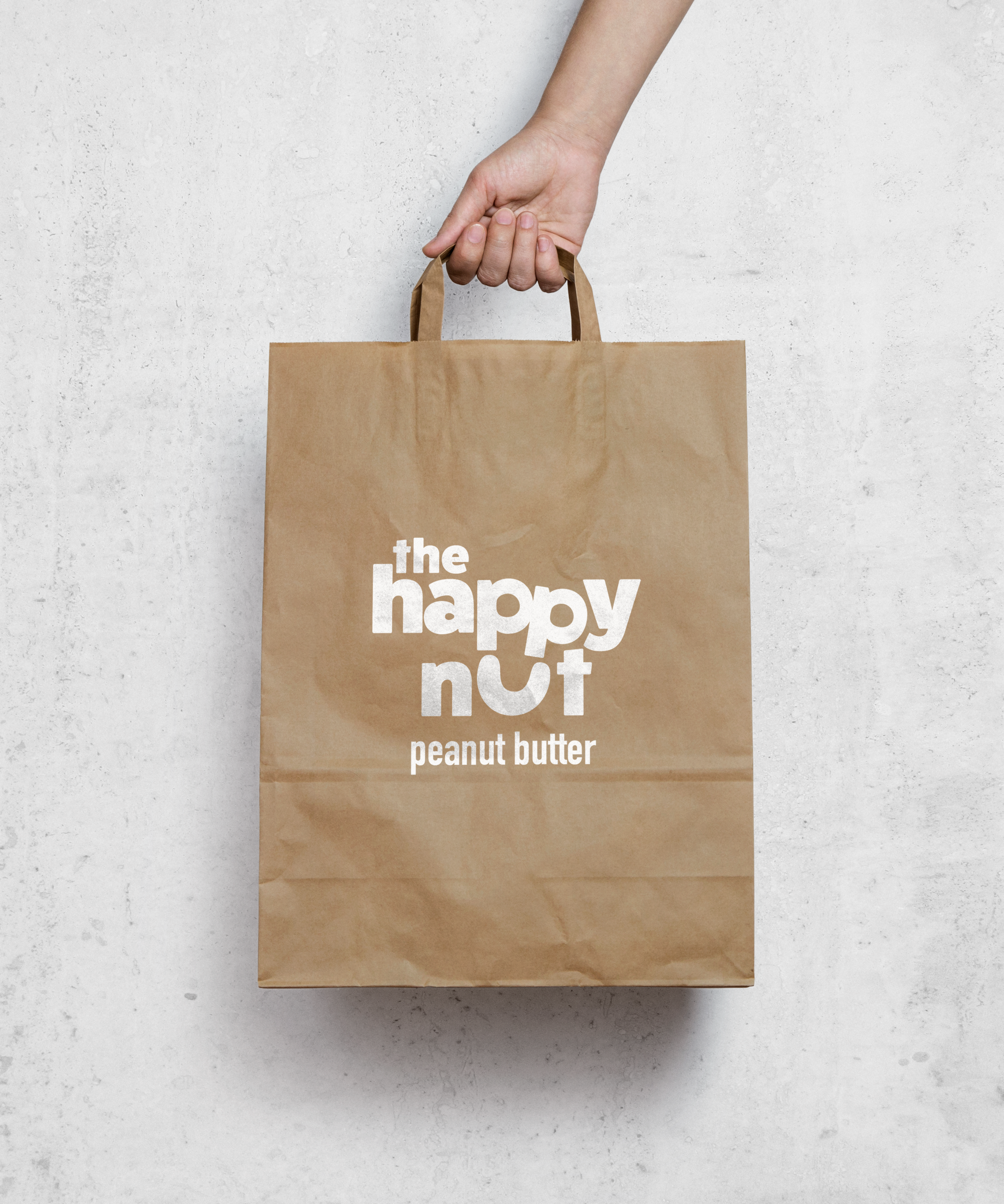 Brown paper bag with white text reading "the happy nut peanut butter" on a white textured surface.