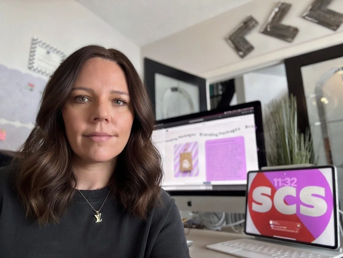 A woman with wavy brown hair taking a selfie in an office or workspace. Behind her are two screens: a large desktop monitor displaying colorful content, and a tablet on a stand displaying the words "SCS" with a red and purple background.