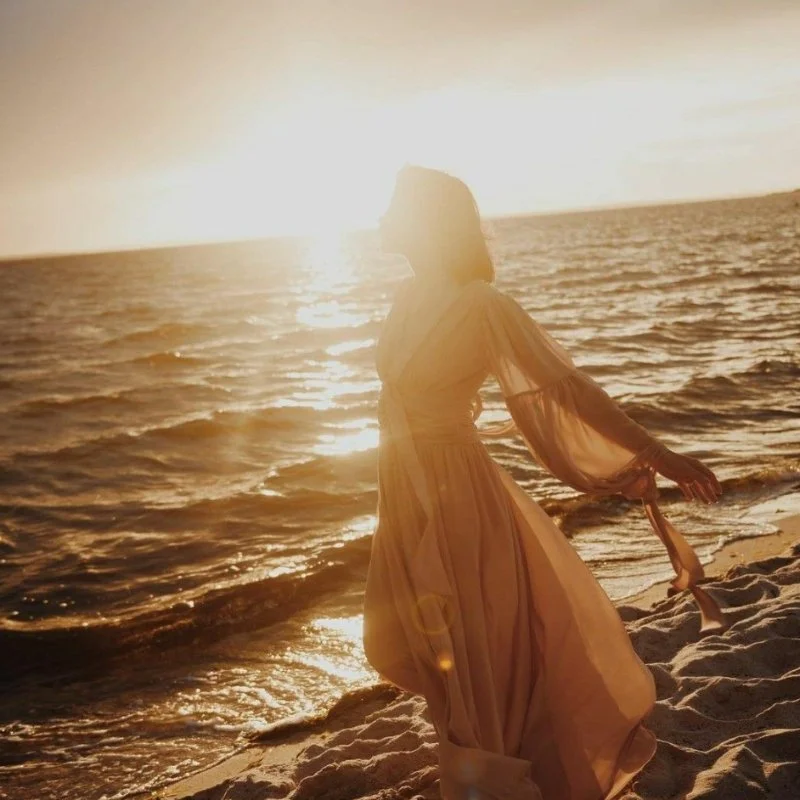 Woman in a flowing dress standing open-armed at the shore at sunset, finally settling after living in survival mode without realizing it