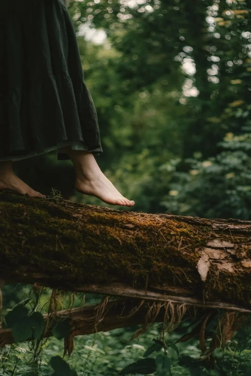 A woman in a long dark skirt stands barefoot on a mossy fallen log in a green forest, a calm moment for anyone asking, do you need to have experienced trauma to explore somatic work?