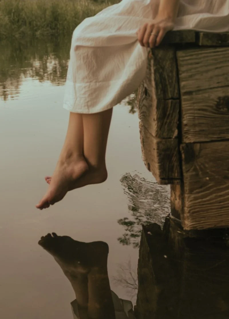 Woman sitting barefoot on a wooden dock with her feet resting above calm water.