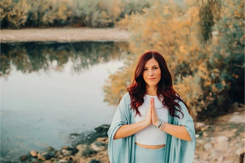 A woman with long red hair and a soft blue wrap stands by a calm river with her hands pressed together, a peaceful pause from holiday stress and your nervous system.