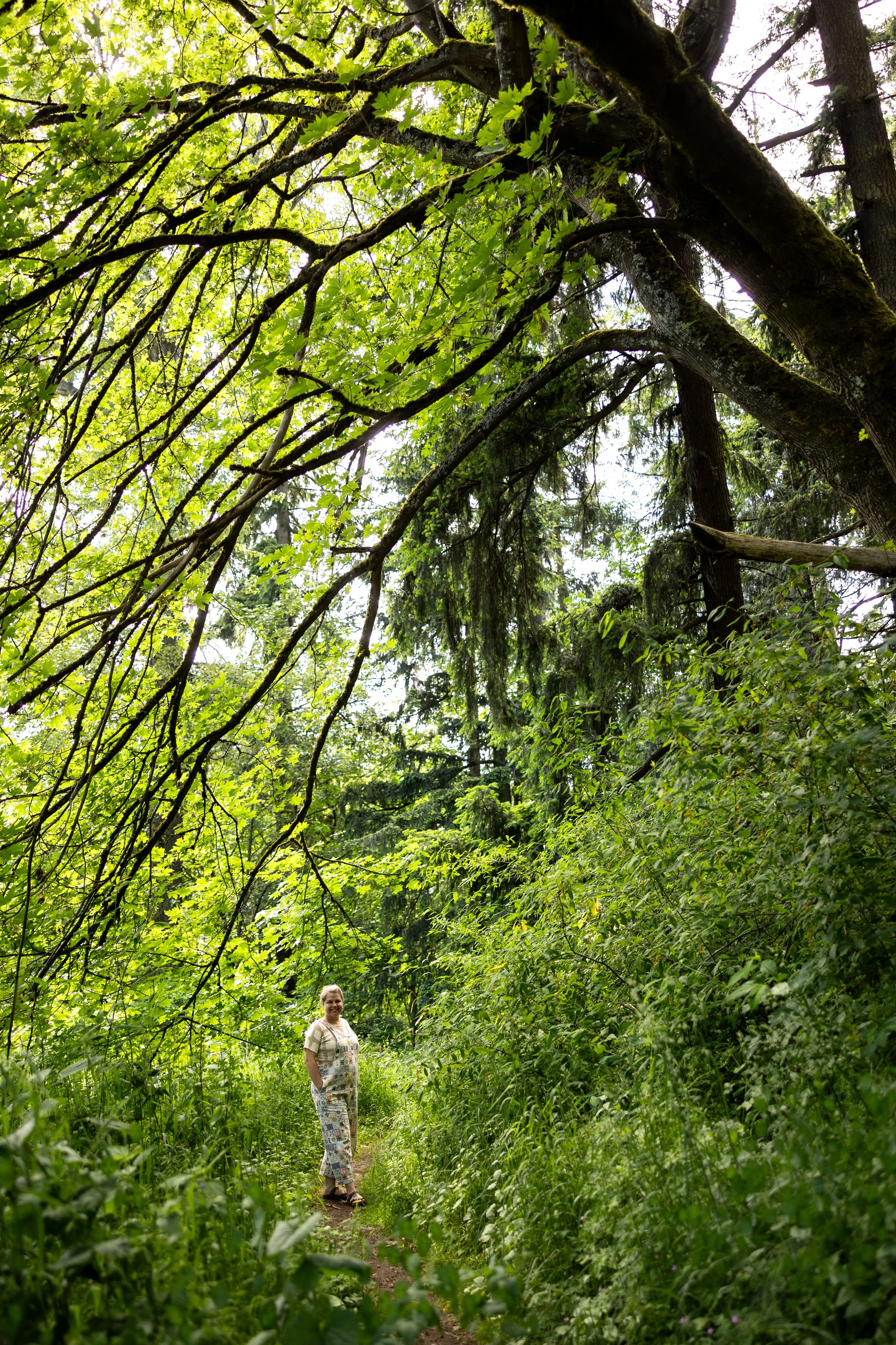 A woman standing on a narrow forest trail surrounded by lush green trees and foliage, with sunlight filtering through the leaves.