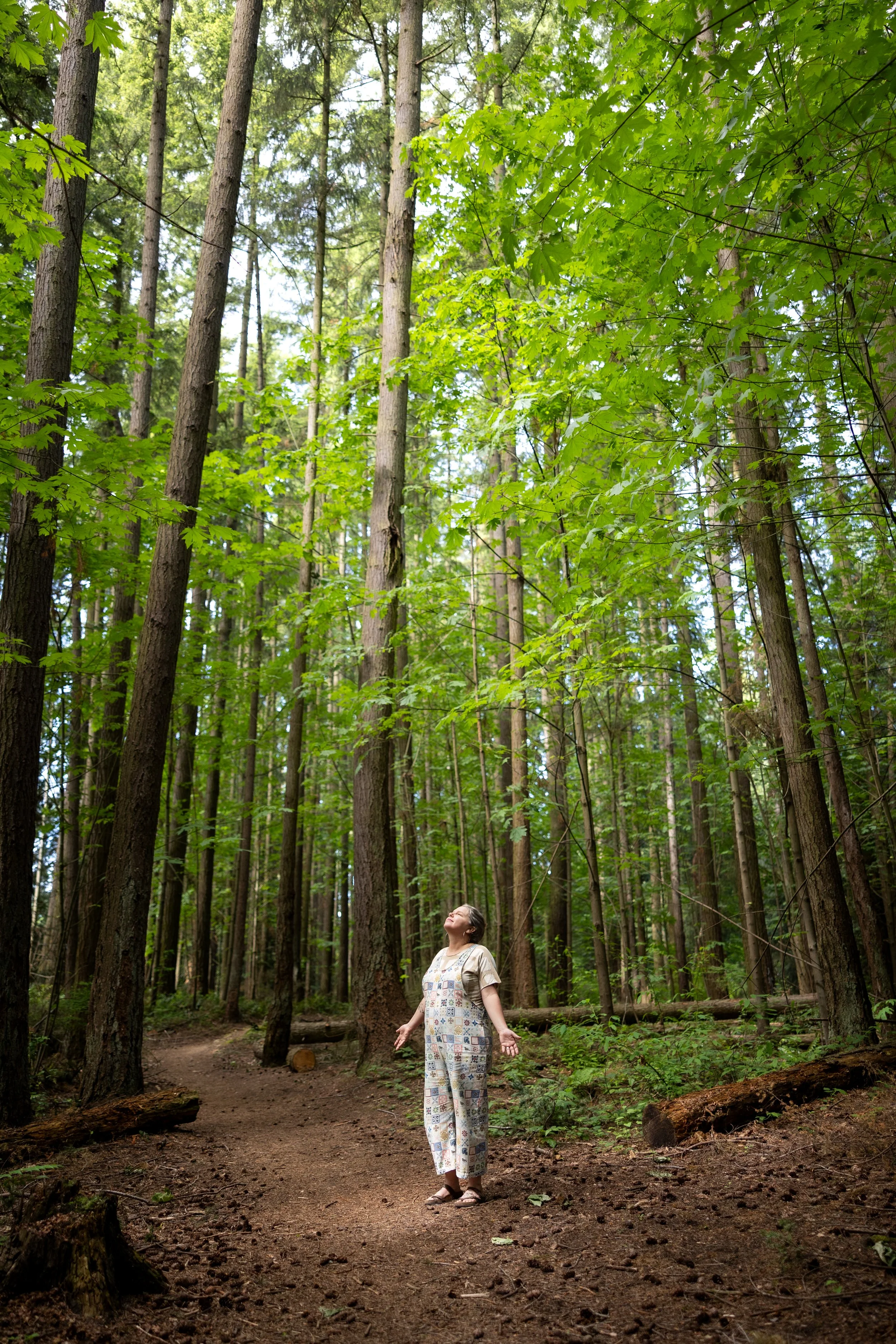 Virginia in a patterned jumpsuit  with her arms down and looking up, surrounded by maple and pine trees. r hands down