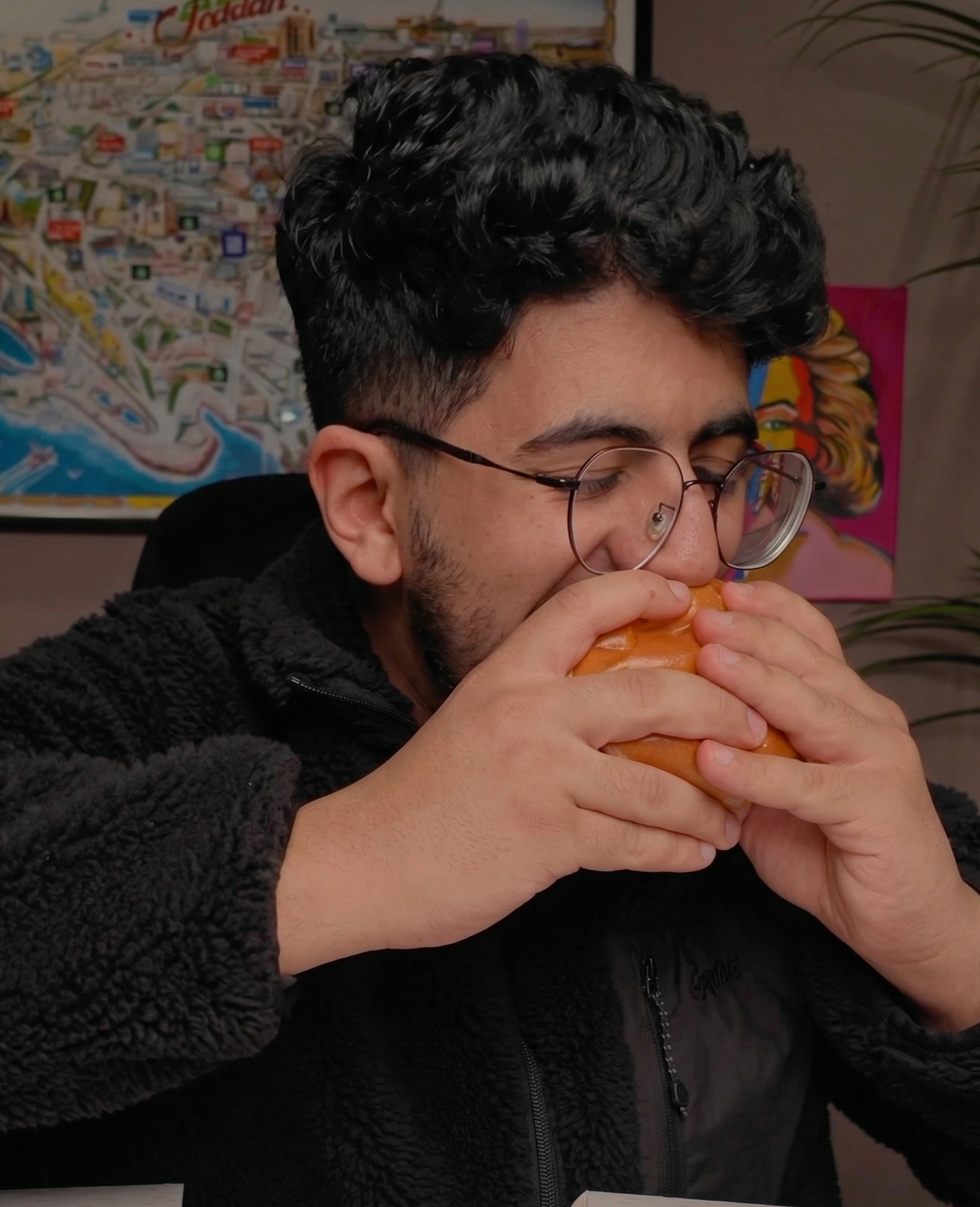 Young man wearing a red and white keffiyeh and glasses, smiling, in front of a blurred background.
