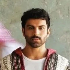 A man with dark hair and a beard standing with his hands in his pockets, wearing a white shirt and a colorful patterned jacket, flanked by two men in white traditional Middle Eastern thobes with red and white checkered head coverings, walking past him in a room with a green wall and framed pictures.