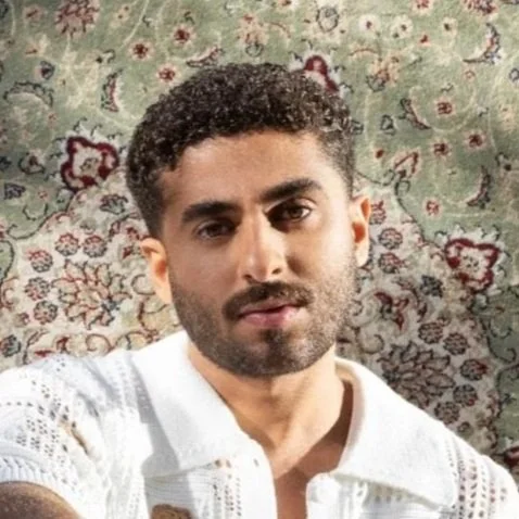 A man with curly dark hair and a beard lying on a patterned rug, wearing a white textured shirt, looking at the camera with a slight smile.