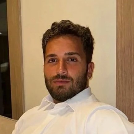 A man with a beard and curly hair dressed in a white traditional outfit, sitting on a beige chair in a modern indoor setting with wooden and glass decor.