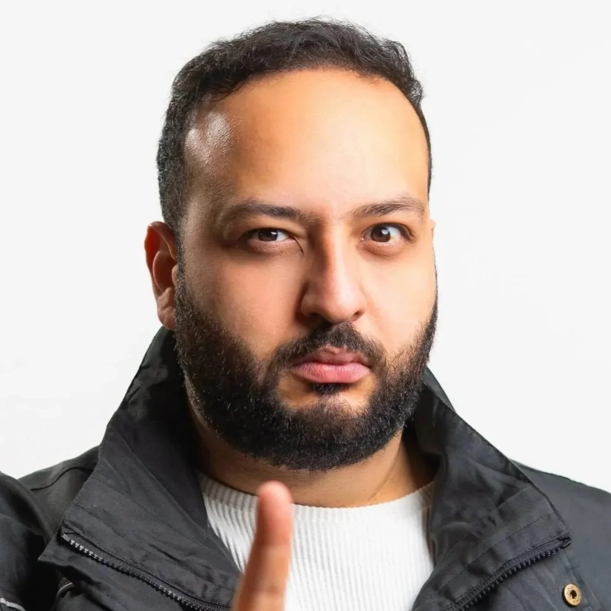 Man with dark hair and beard wearing a black jacket, holding up one finger.