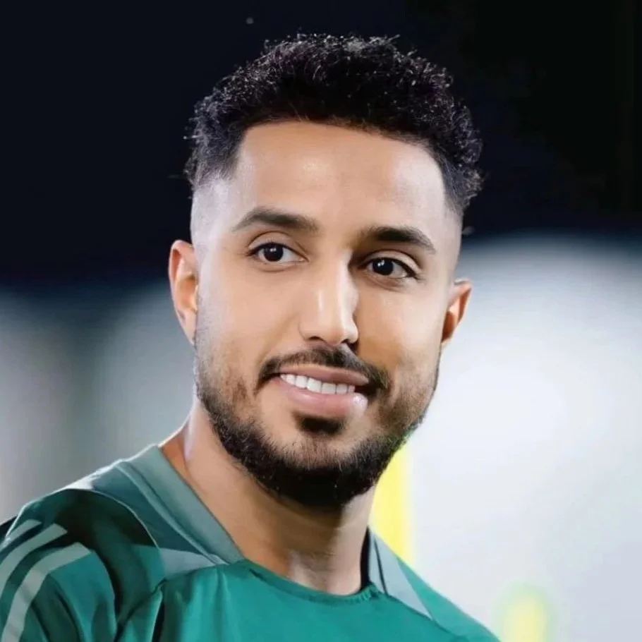 A man with short curly black hair and a beard, wearing a green sports jersey with Saudi Arabia's emblem, standing on a sports field.