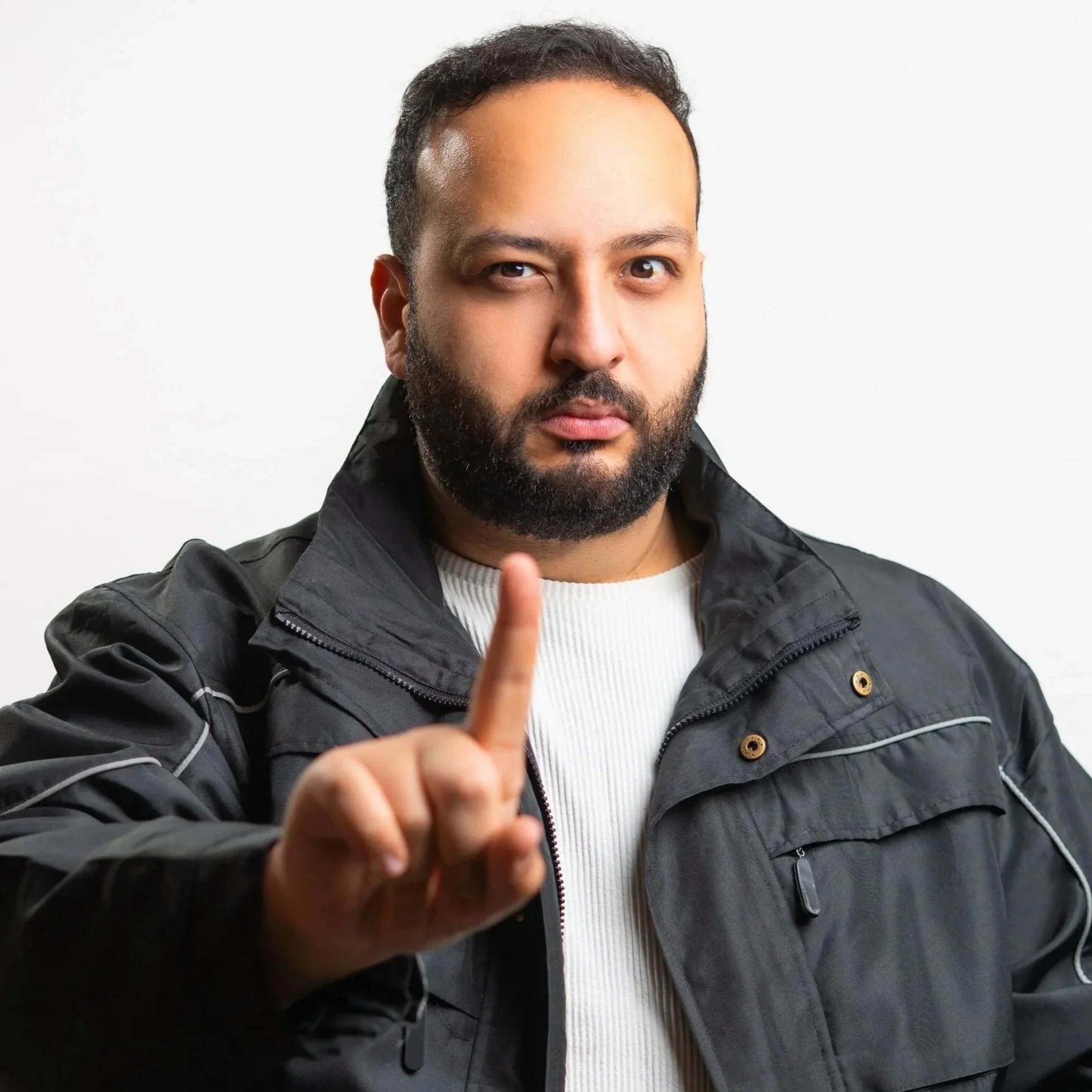 Man with dark hair and beard wearing a black jacket, holding up one finger.