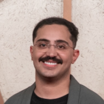 A man with glasses and a mustache smiling, wearing a dark shirt and gray jacket, standing in front of a textured wall background.