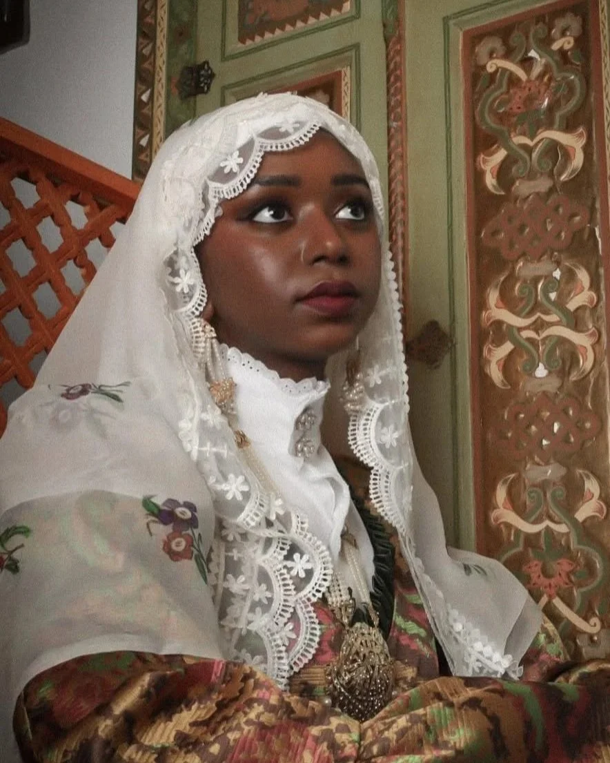 A woman dressed in traditional clothing, wearing a white lace head covering and ornate jewelry, sitting in front of a decorated, painted wall with intricate designs.