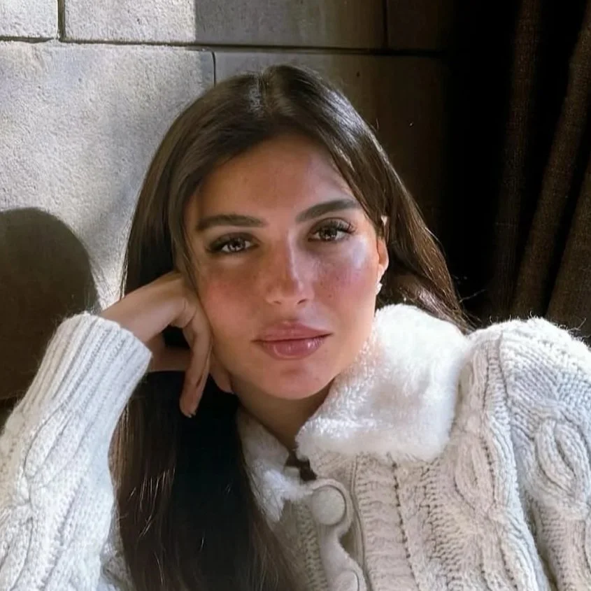 A woman with long dark hair, wearing a cream-colored cable knit sweater, is sitting next to a stone wall and a dark curtain. She has her head resting on her hand and is looking at the camera.