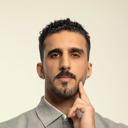 A man with dark curly hair, a beard, and a mustache, dressed in a light gray suit, is standing with one arm crossed and the other hand touching his chin, looking thoughtful.