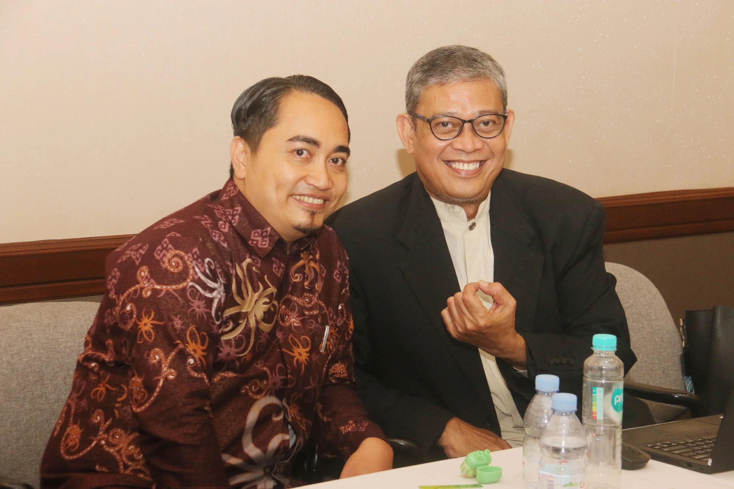 Two men are sitting at a table smiling, with water bottles and hand sanitizer on the table. One man is wearing glasses and a patterned dark maroon shirt, while the other is wearing a black blazer and a white shirt.