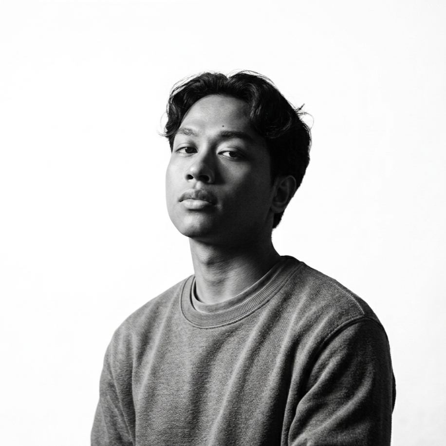 Black and white portrait of a young man with wavy hair wearing a sweatshirt, looking at the camera with a neutral expression against a plain light background.