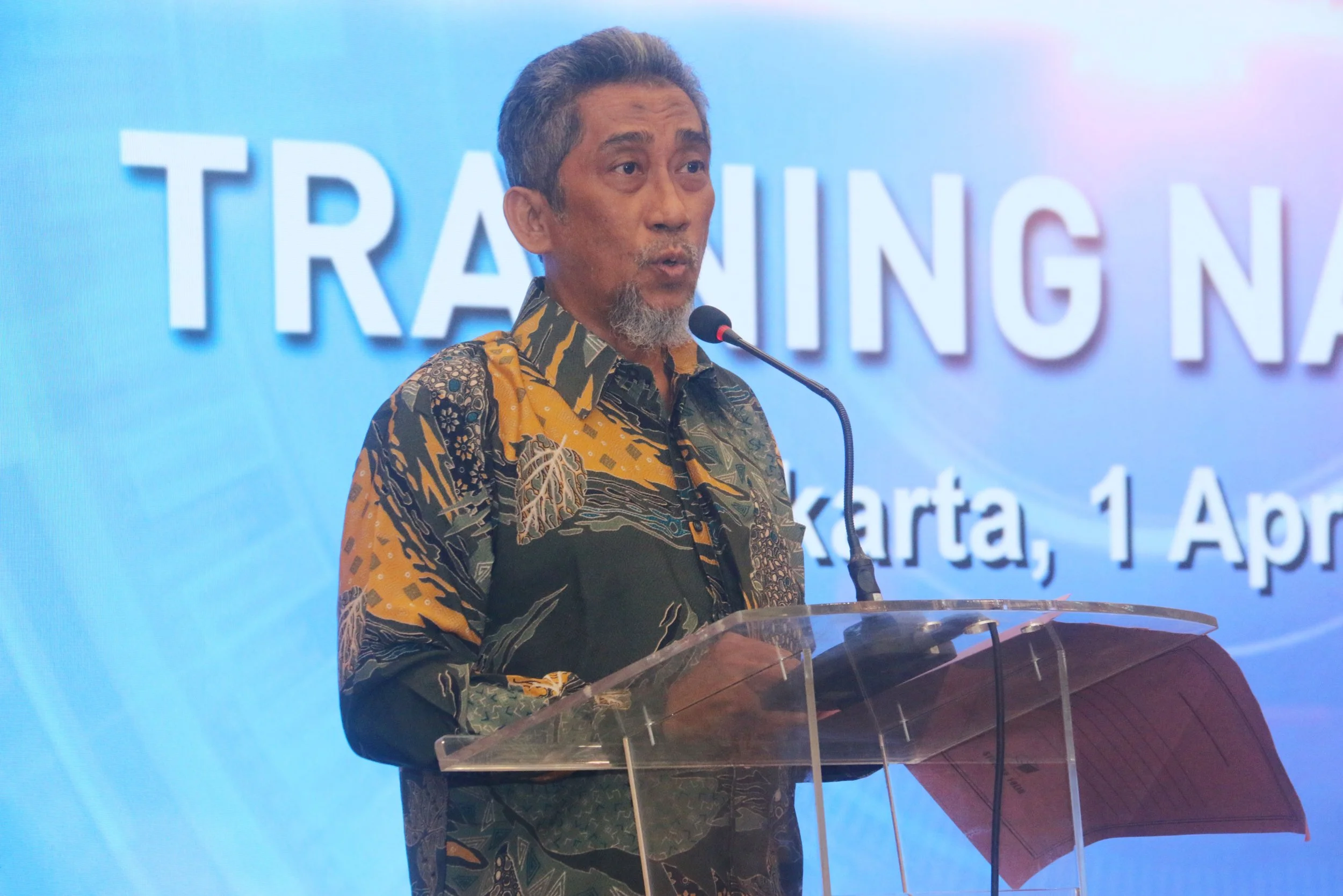 A man with gray hair and a beard speaking at a podium during a conference or event. He is wearing a yellow and green batik shirt. In the background, a large screen displays the partially visible word 'TRAINING' and some text about Jakarta, Indonesia.
