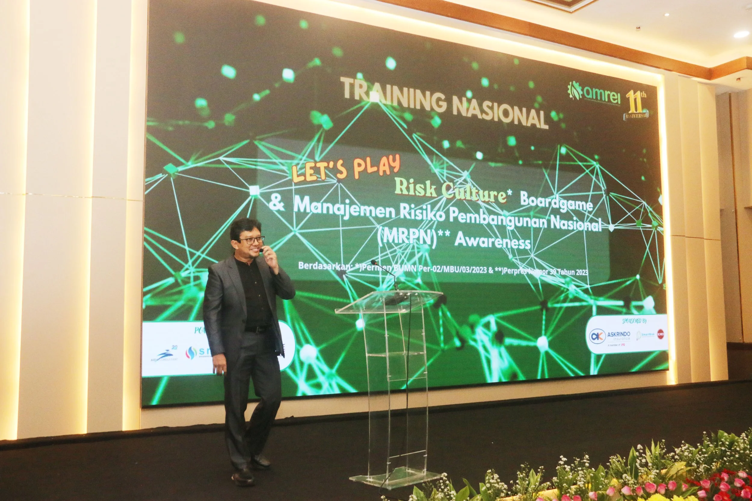 A man in a black suit is standing on a stage with a microphone in hand in front of a large screen. The screen displays the text 'Training Nasional' and 'Let's Play Risk Culture,' along with graphics of interconnected green dots and lines. There are logos for sponsors and organizers at the bottom of the screen, and a flower arrangement is visible in the foreground.