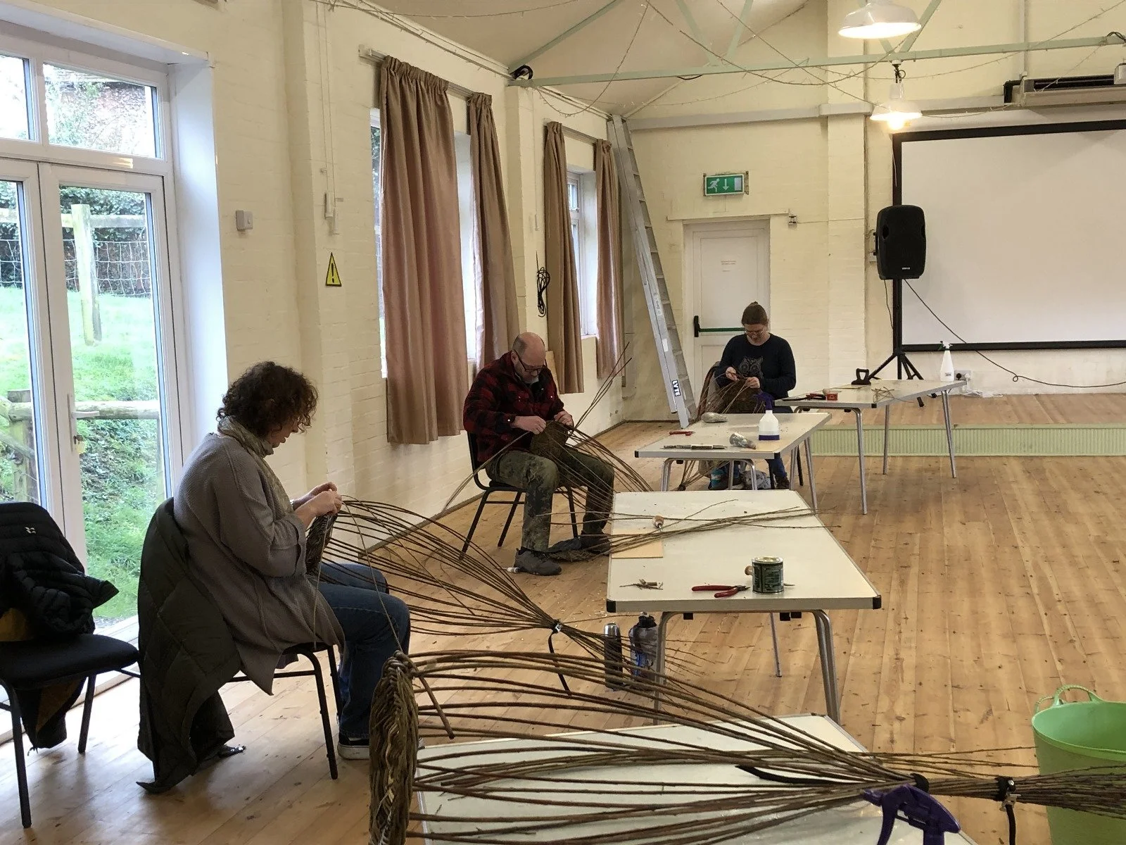 Four people working together on basket weaving projects in a well-lit, spacious room with white brick walls and wooden floors. Tables with tools and materials are in front of them, and a ladder leans against the wall.