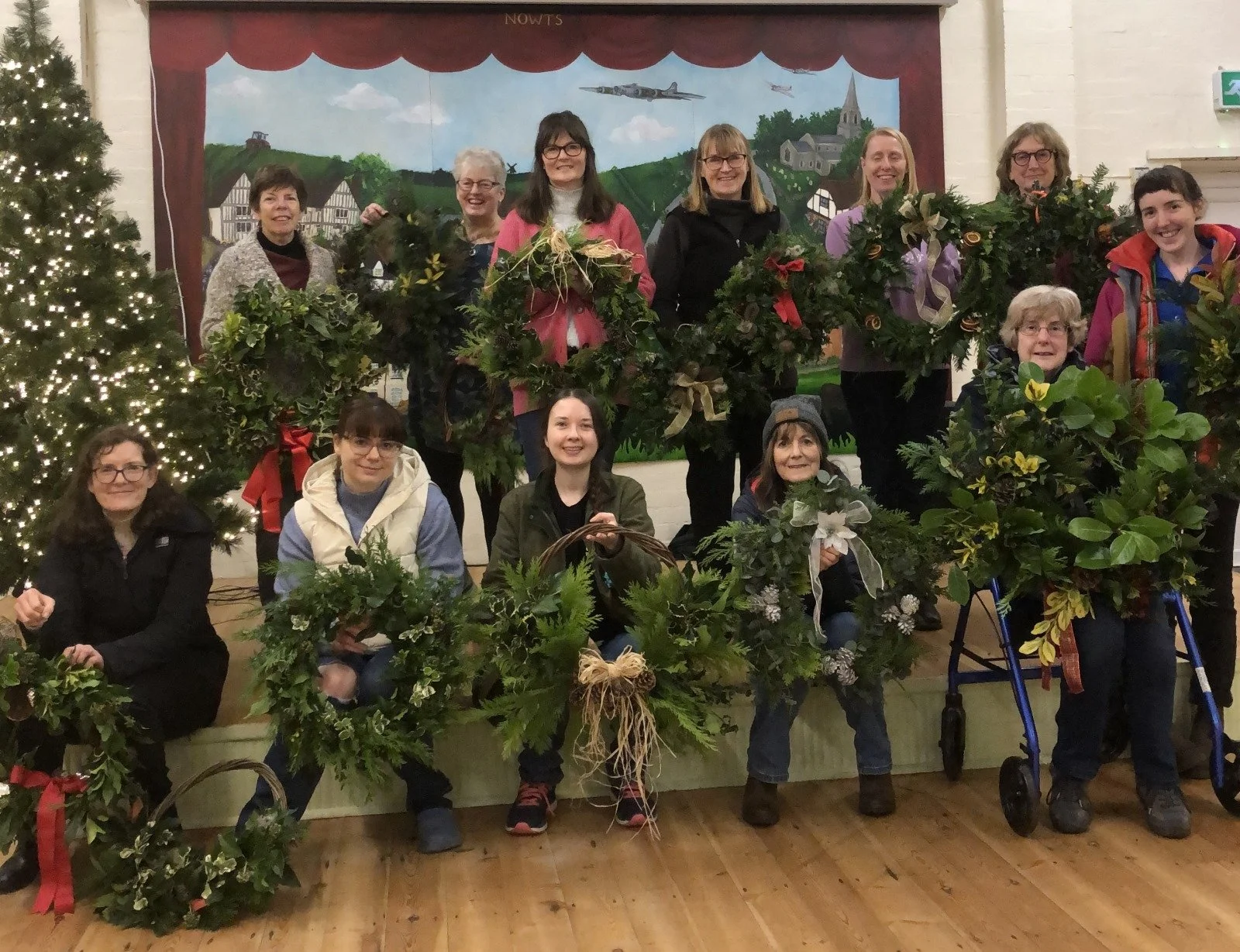 Group of women holding holiday wreaths indoors, decorated Christmas tree on the left, and a festive backdrop with trees and a church in the background.
