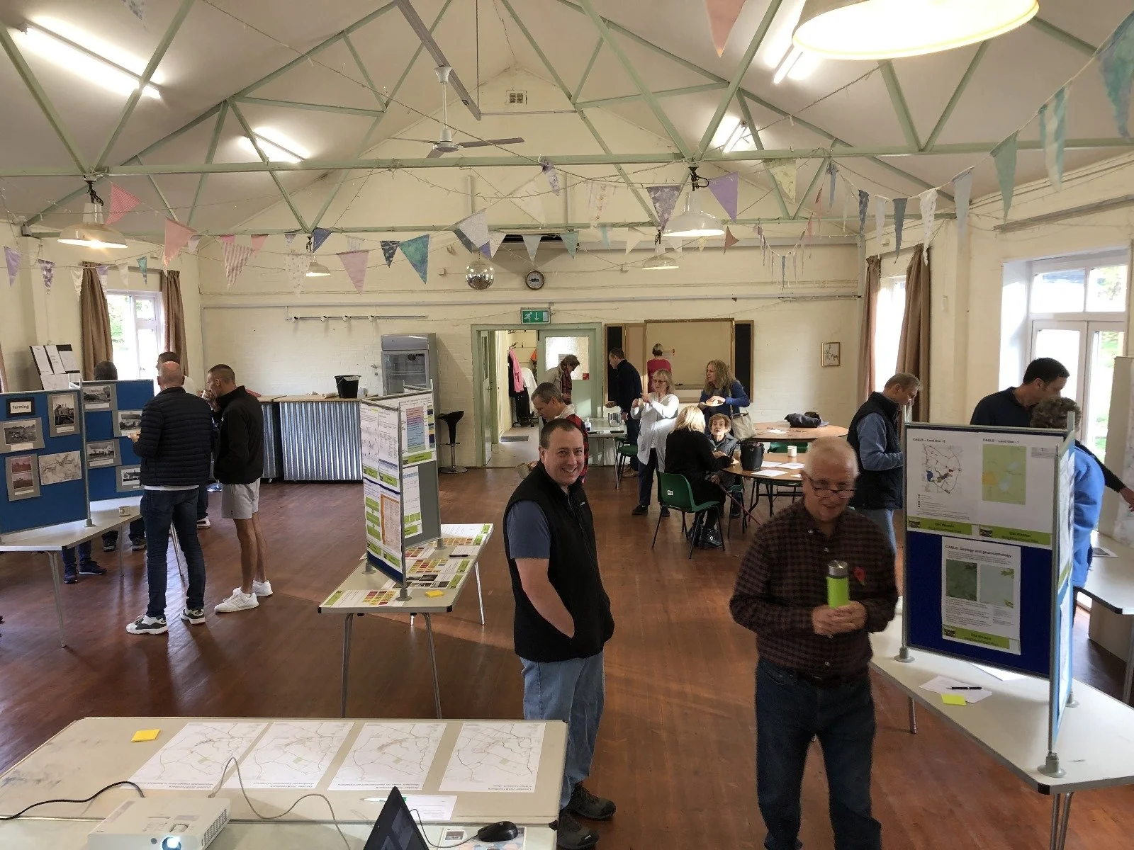 Community event in a large hall with display boards, tables, and people interacting; decorated with colorful bunting flags hanging from the ceiling.