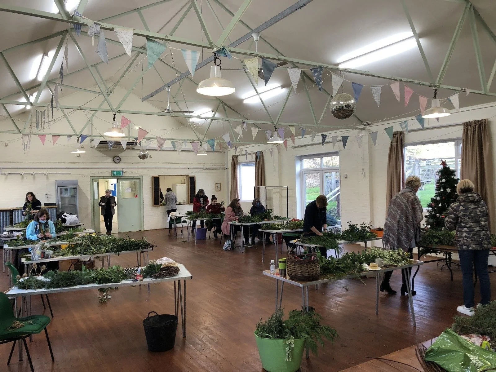 People decorating tables with greenery and Christmas decorations in a decorated community hall with pastel bunting, a Christmas tree, and large windows.