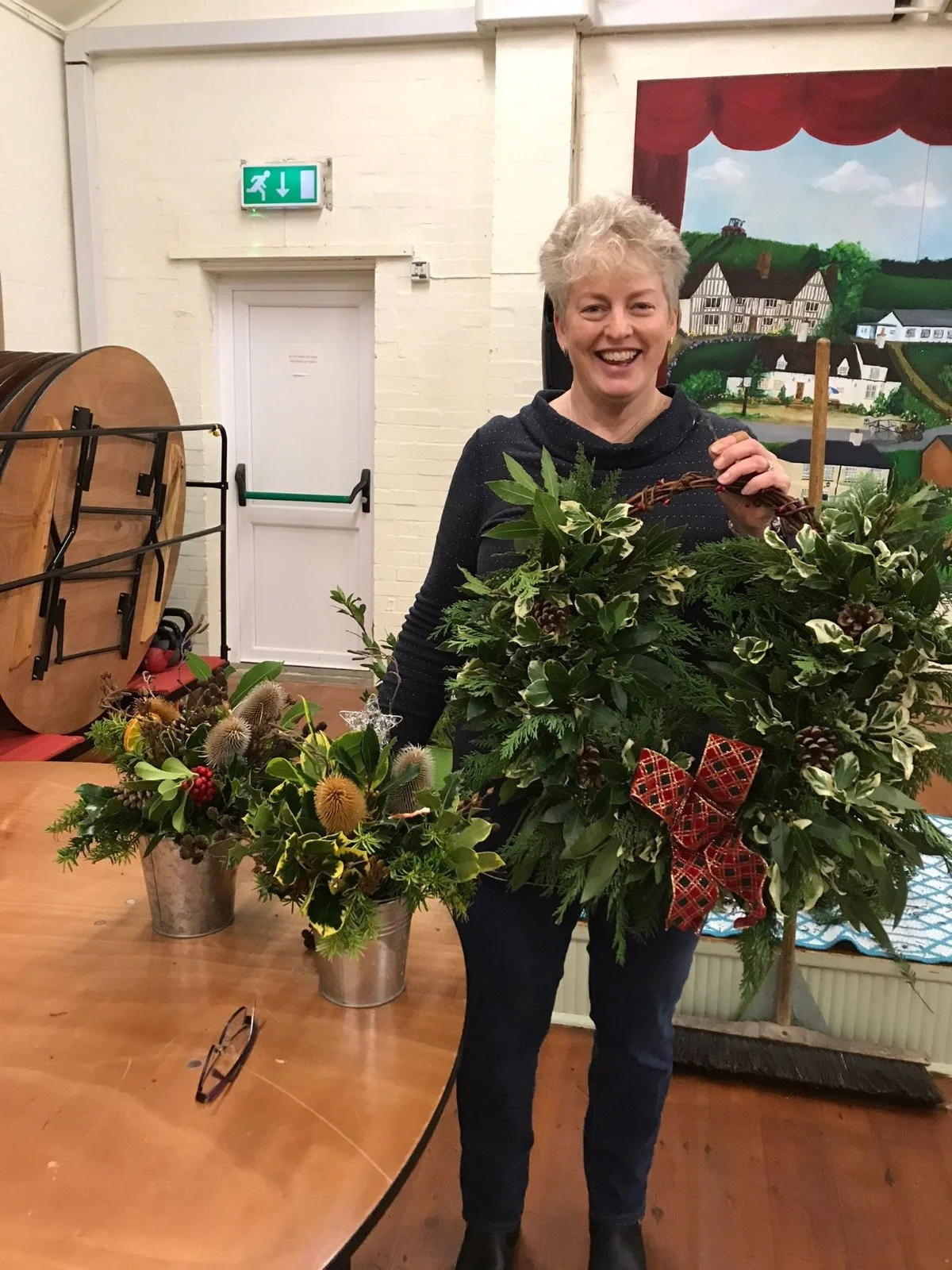 A woman with short blonde hair holding a large Christmas wreath decorated with pinecones, holly, and a red bow, smiling inside a room with a table and floral arrangements.