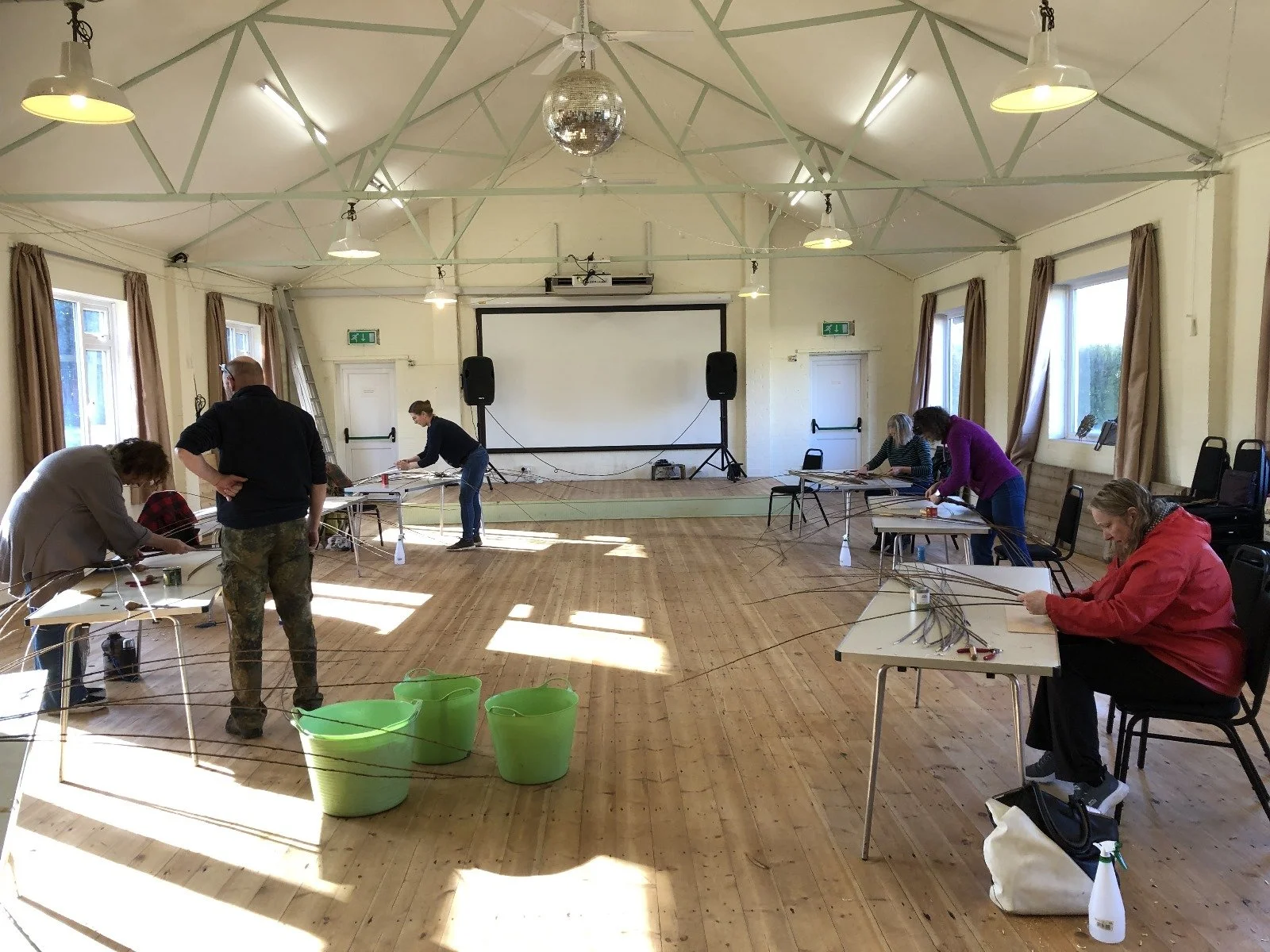 Group of people working on craft or wire projects at tables in a spacious room with wooden floor and large windows, some connecting wires and tools on the tables.