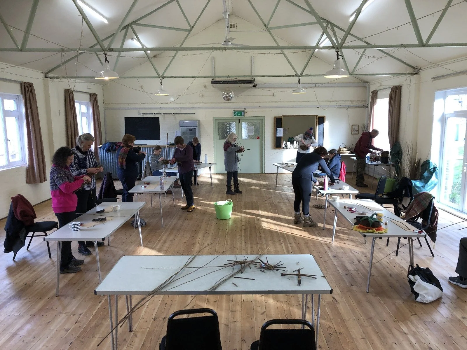 A group of people in a spacious room participating in a craft workshop, with tables arranged with tools and materials, and windows letting in natural light.