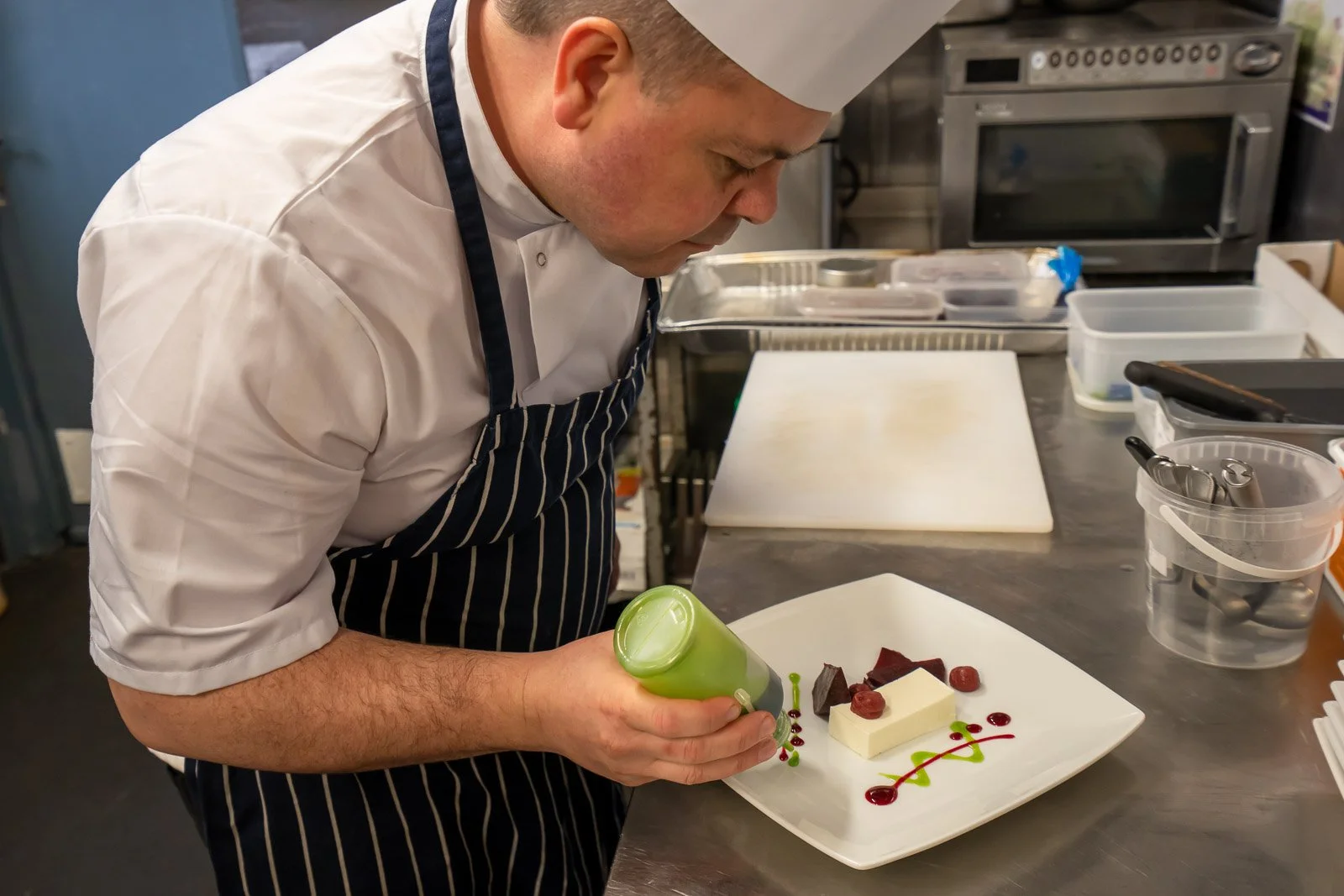 Chef decorating a dessert with green sauce on a white plate in a professional kitchen.