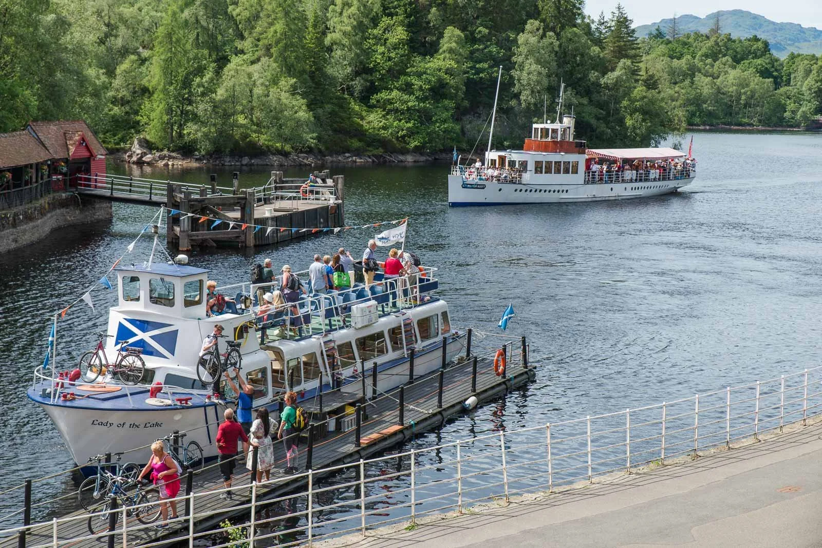 People boarding a boat at a dock on a lake, with lush green trees and hills in the background.