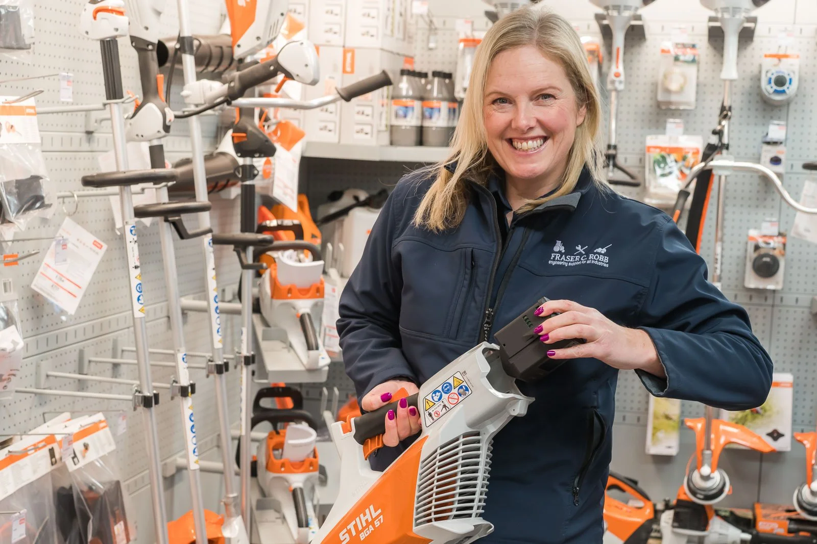 A woman with blonde hair smiling in a hardware store, holding a leaf blower with the brand STIHL, surrounded by gardening tools and equipment on shelves.