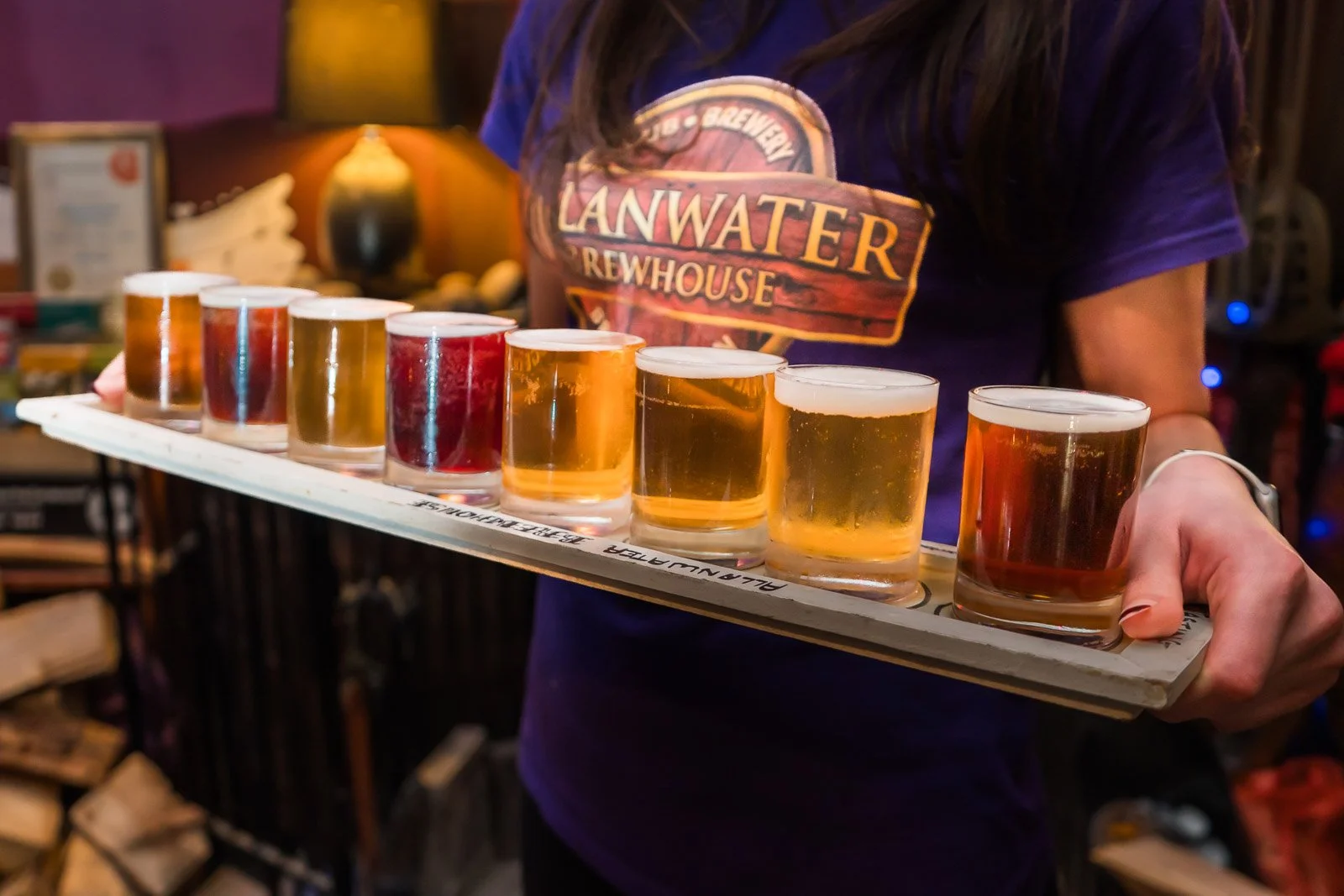 A person holding a wooden flight with nine small beer glasses, each filled with different types of beer, at Allanwater Brewhouse in Bridge of Allan