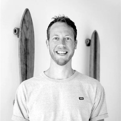 Black and white portrait of a smiling man wearing a light-colored t-shirt, standing in front of two vertically mounted longboards on a wall.