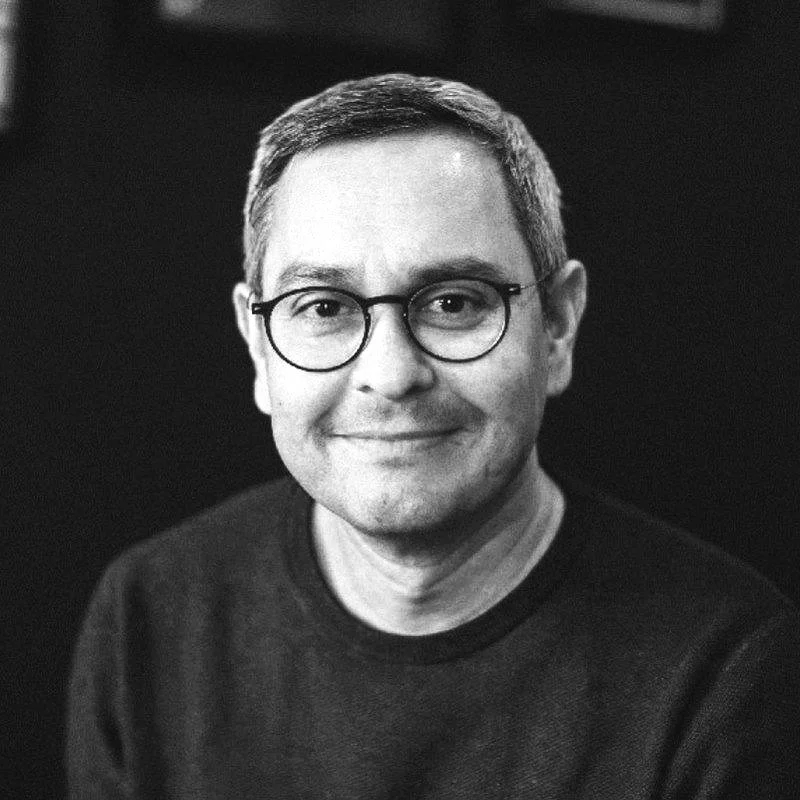 Black and white portrait of a man with short hair, glasses, and a slight smile, wearing a dark sweater against a dark background.